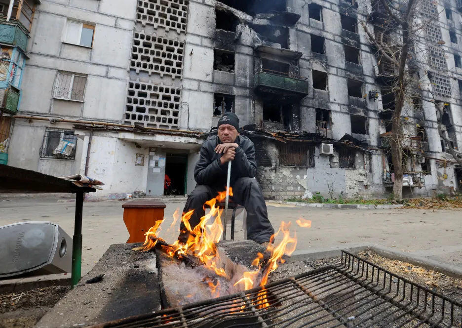 Obyvatel Mariupolu sedí u ohně před těžce poškozeným bytovým domem